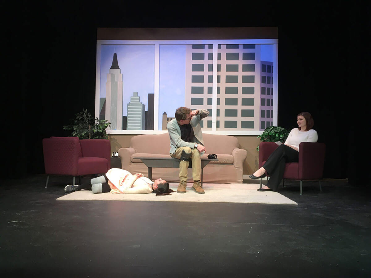 One actor looks at another sitting on a couch and turned away from a woman passed out on the floor