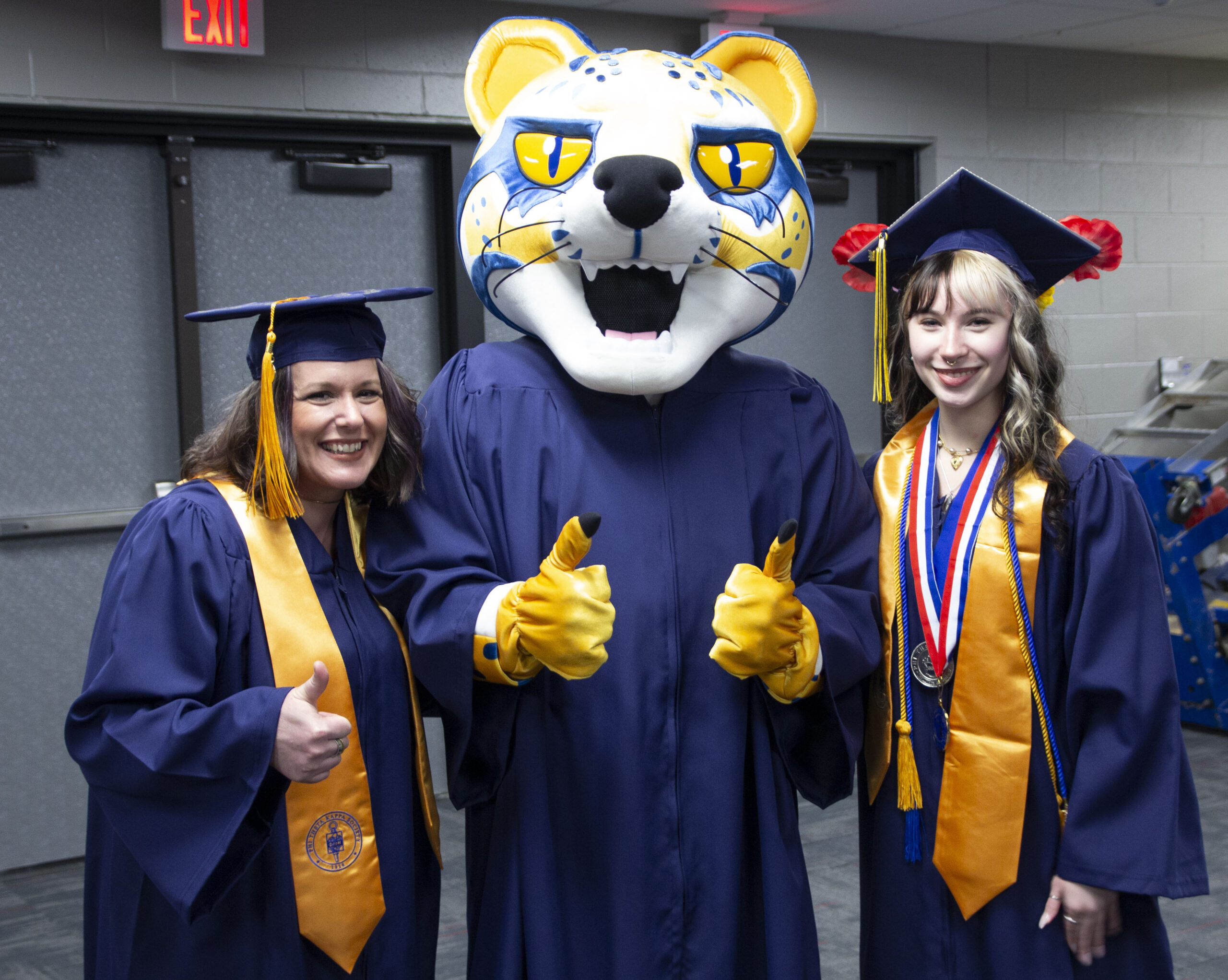 two students in regalia smiling with the ocelot mascot